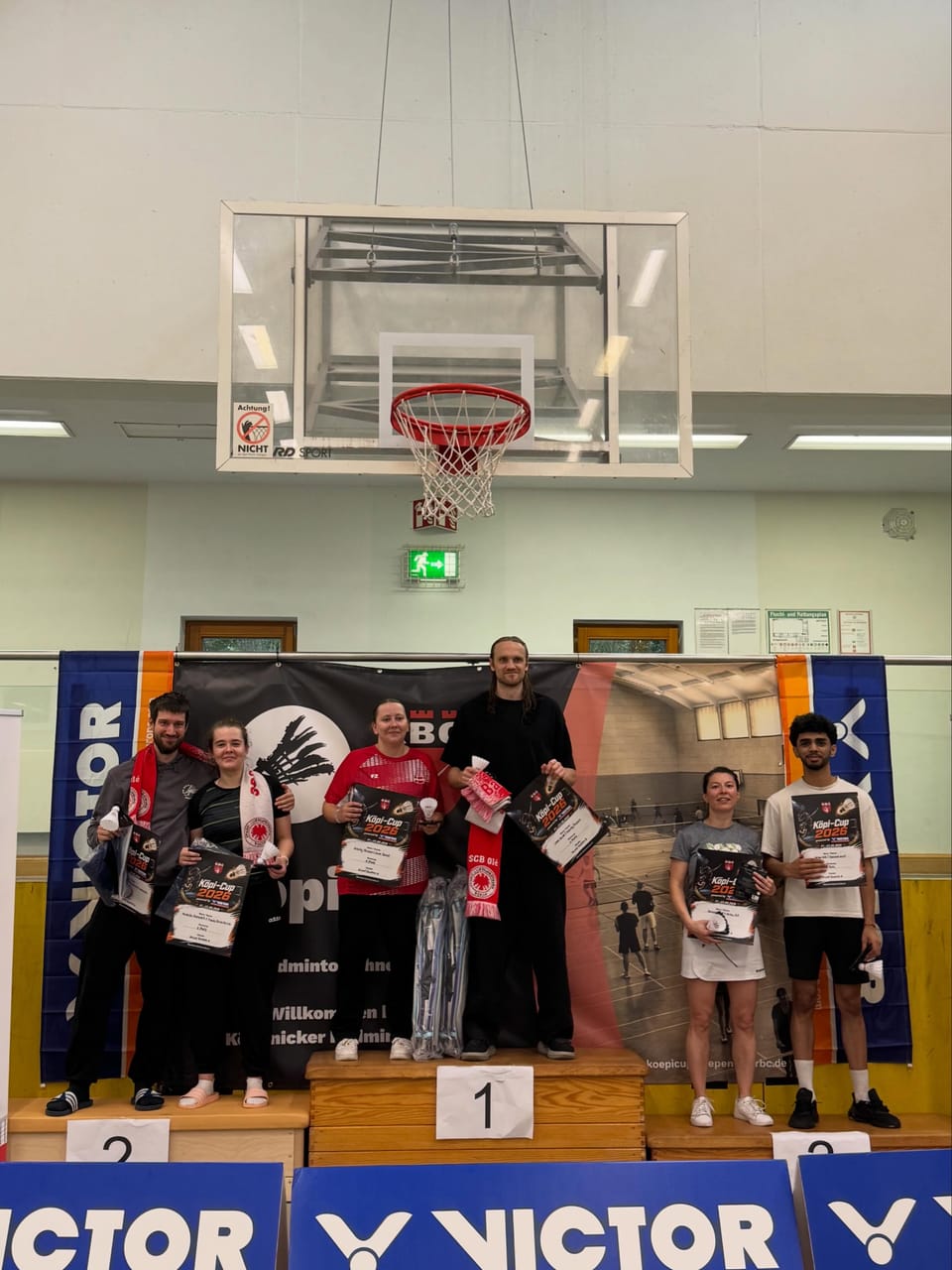 Drei Mixed-Paare stehen mit Urkunden, Bällen und Schals auf dem Siegerpodest in einer Sporthalle unter einem Basketballkorb.